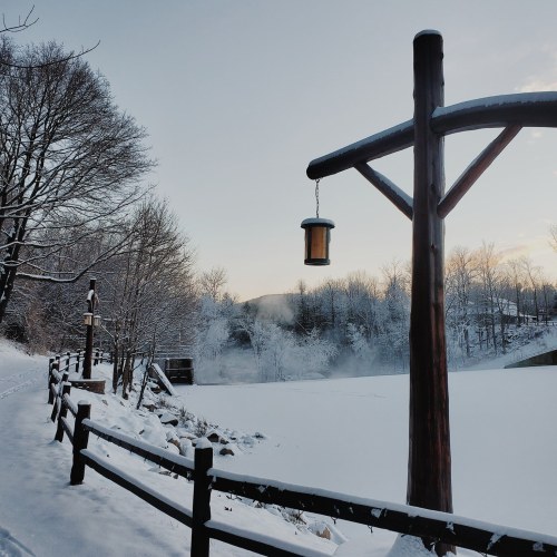 A snowy landscape with a wooden post and lantern, surrounded by trees and a frozen lake under a clear sky.