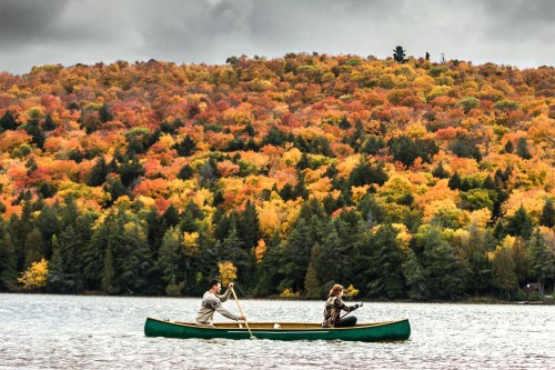 Two people paddle a canoe on a lake surrounded by vibrant autumn foliage and a cloudy sky.