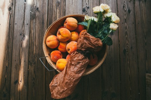 A wooden basket filled with peaches and a bouquet of white roses wrapped in brown paper on a wooden surface.