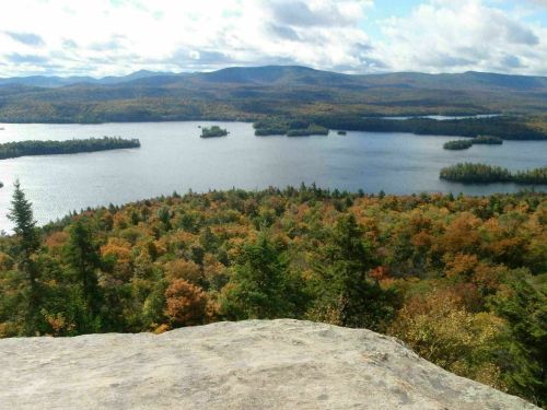 A scenic view of a lake surrounded by colorful autumn foliage and distant mountains under a partly cloudy sky.