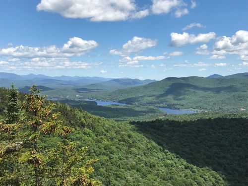 A panoramic view of lush green mountains and a serene lake under a blue sky with fluffy white clouds.