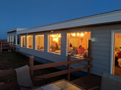 A cozy restaurant at dusk, with warm lights glowing from large windows and diners enjoying their meals inside.