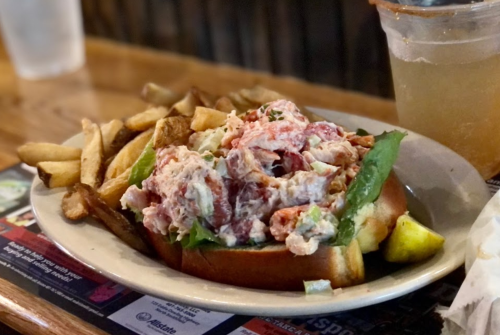 A lobster roll topped with creamy lobster salad on a bun, served with fries and a drink in the background.