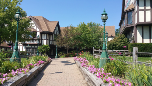 A charming pathway lined with flowers, leading to Tudor-style buildings under a clear blue sky.