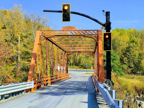 A bright orange metal bridge with a traffic light, surrounded by autumn foliage and a clear blue sky.
