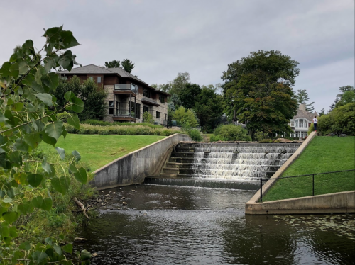 A serene scene featuring a waterfall cascading into a calm river, surrounded by greenery and residential buildings.