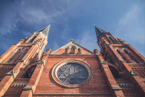A low-angle view of a brick church with two tall spires and a large circular window against a blue sky.