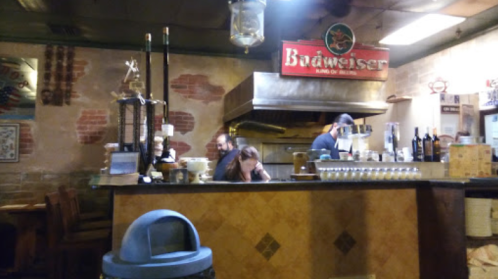 A cozy bar scene with a Budweiser sign, featuring a bartender and patrons at the counter in a rustic setting.