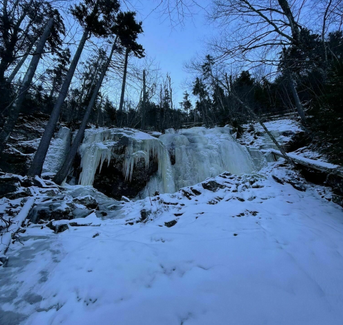A frozen waterfall surrounded by snow-covered rocks and trees, under a twilight sky.