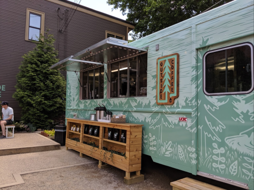 A green coffee truck with a wooden serving counter, surrounded by greenery and a person sitting nearby.