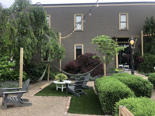 A cozy outdoor space with hammocks, seating, and greenery, illuminated by string lights against a rustic building.