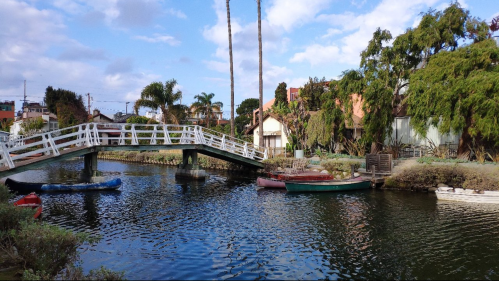 A serene canal scene featuring a white bridge, palm trees, and colorful boats moored along the water's edge.