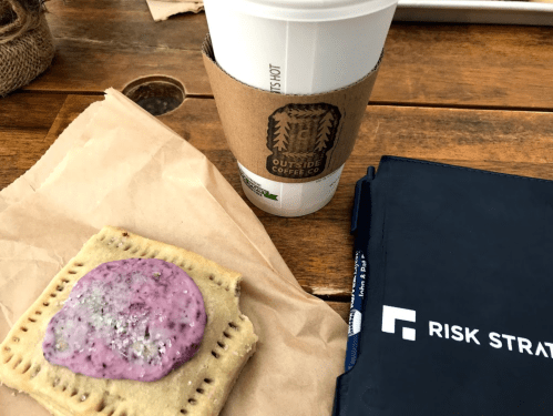 A coffee cup and a frosted pastry on a brown paper napkin, with a notebook in the background on a wooden table.