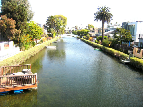 A serene canal lined with palm trees and houses, featuring small boats and lush greenery under a clear blue sky.