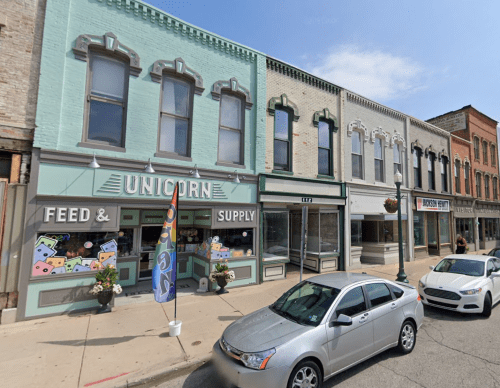A street view of colorful storefronts, including a shop named "Unicorn Feed & Supply," with parked cars in front.