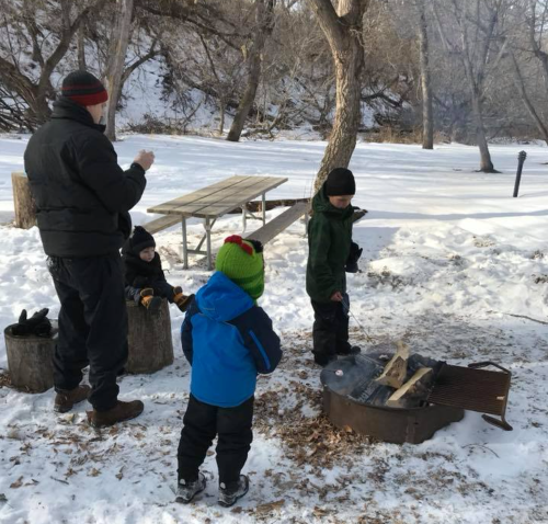 A man and two children gather around a fire pit in a snowy park, with trees and a picnic table in the background.