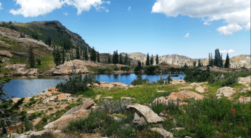 A serene mountain landscape featuring a clear lake, rocky terrain, and lush greenery under a blue sky with clouds.