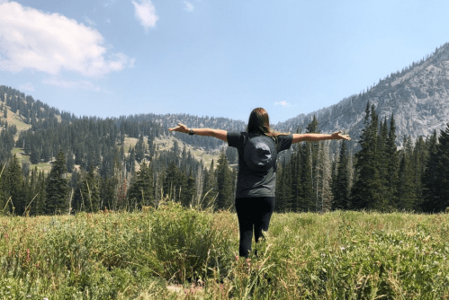 A person stands in a grassy field with arms outstretched, surrounded by mountains and trees under a blue sky.