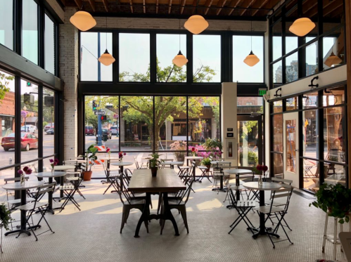 Bright café interior with tables and chairs, large windows, and plants, offering a view of the street outside.