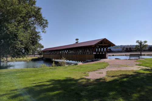 A wooden covered bridge spans a calm river, surrounded by green grass and trees under a clear blue sky.