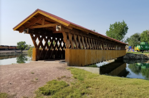 A wooden covered bridge with a red roof spans a calm waterway, surrounded by greenery and a playground in the background.