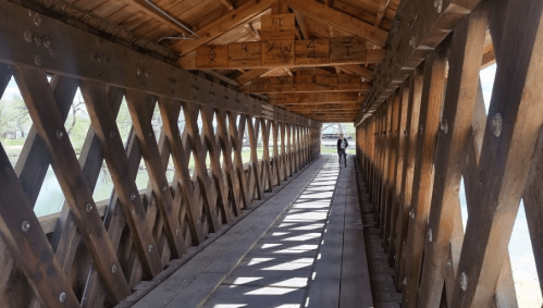A wooden covered bridge interior with diagonal beams and sunlight casting shadows on the floor. A person stands in the distance.
