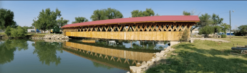 A wooden covered bridge with a red roof spans a calm river, surrounded by greenery and a clear blue sky.