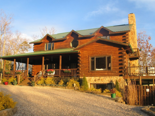 A large log cabin with a green roof, surrounded by trees and a gravel driveway, under a clear blue sky.