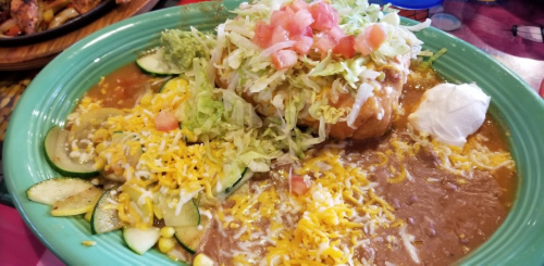 A colorful plate of Mexican food featuring a large burrito topped with lettuce, tomatoes, cheese, and sides of beans and zucchini.