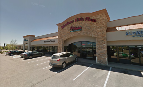 Exterior view of a shopping plaza with stone accents, featuring several storefronts and parked cars in the lot.
