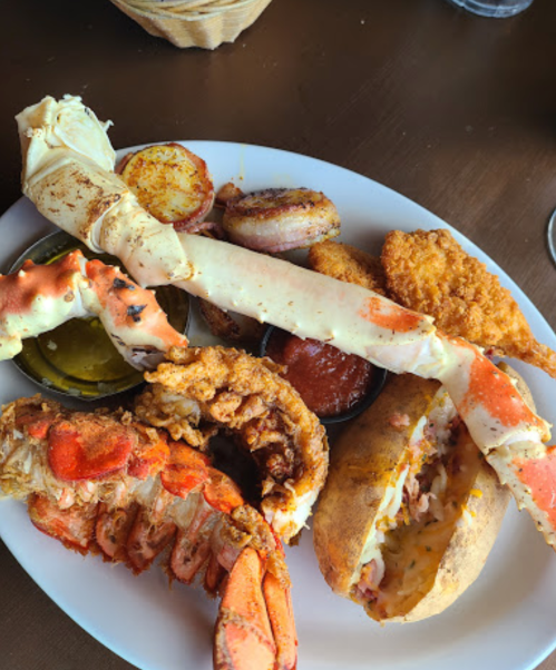 A plate of seafood featuring crab legs, fried shrimp, lobster, and a baked potato with dipping sauces.