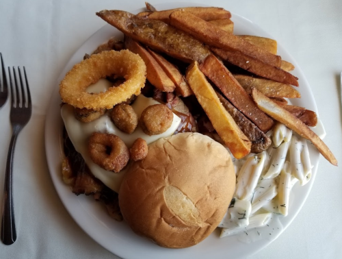 A plate of food featuring a sandwich, onion rings, fried potatoes, and pasta on a white tablecloth.