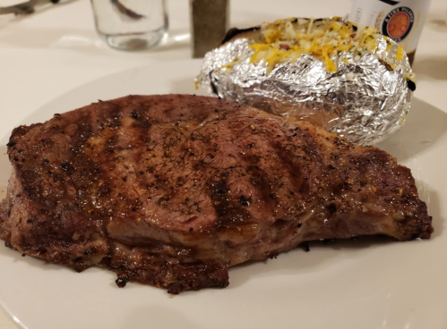 A grilled steak with a perfectly seared crust, served alongside a baked potato topped with cheese and foil.