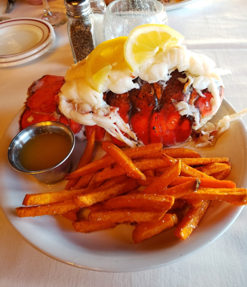 A plate of grilled lobster topped with lemon and onions, served with sweet potato fries and a small cup of sauce.