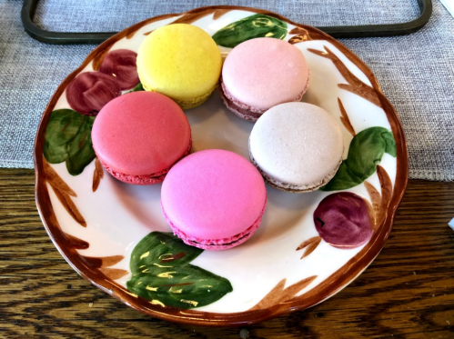 A decorative plate with six colorful macarons: yellow, pink, purple, and red, arranged on a floral-patterned dish.