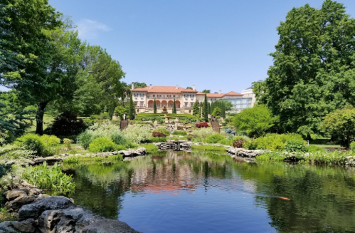 A serene garden scene featuring a pond, lush greenery, and a grand building in the background under a clear blue sky.