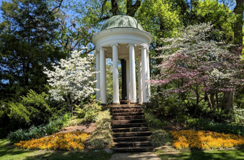 A white gazebo with a green dome surrounded by blooming trees and colorful flowers in a lush garden.