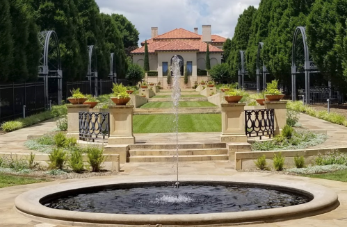 A serene garden with a central fountain, manicured lawns, and elegant landscaping leading to a grand building in the background.