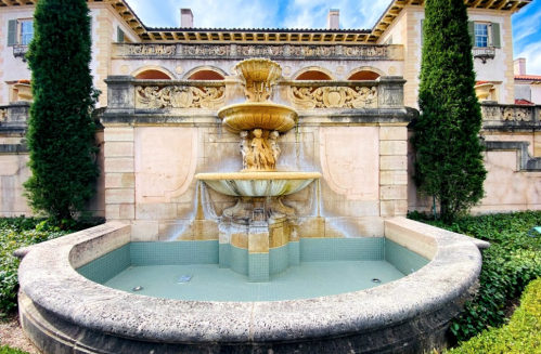A decorative stone fountain with flowing water, surrounded by lush greenery and elegant architecture in the background.