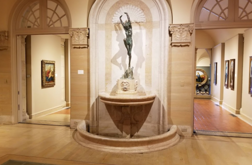 A bronze statue of a dancer stands in a stone fountain, framed by elegant archways in an art gallery.