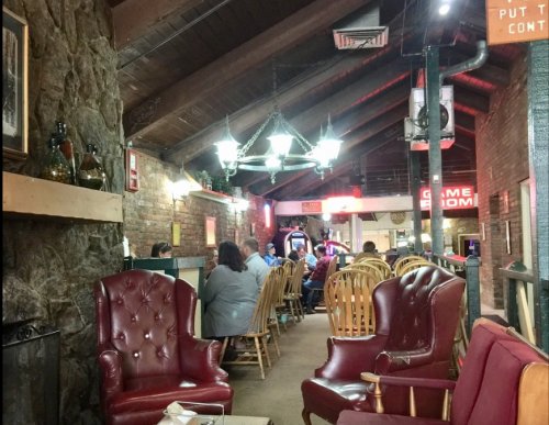 Cozy interior of a rustic restaurant with wooden chairs, stone walls, and a game room in the background.