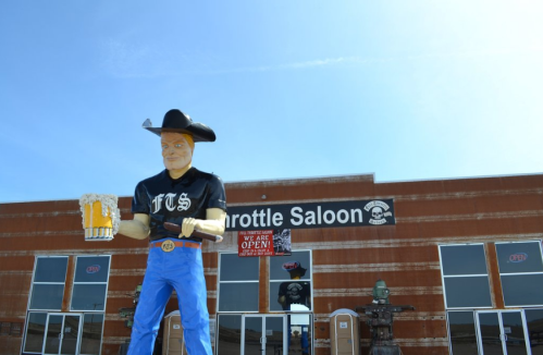 A large cowboy statue holding a beer stands in front of a rustic saloon with an "Open" sign.