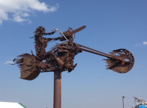 A large metal sculpture of a motorcycle rider, crafted from rusted materials, against a clear blue sky.