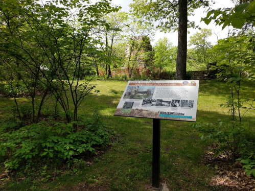 A historical sign in a wooded area, featuring images and text about local history, with ruins visible in the background.