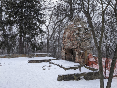 A stone fireplace surrounded by snow-covered ground and trees, with construction barriers in the background.