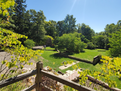 A sunny park scene with green grass, trees, and stone ruins, viewed from a wooden fence. Two people are walking in the distance.