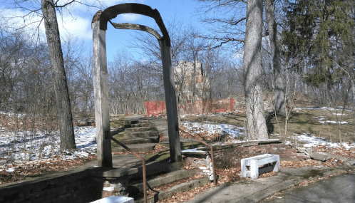 An old, weathered archway leads to a staircase in a wooded area, with remnants of snow and a distant building.