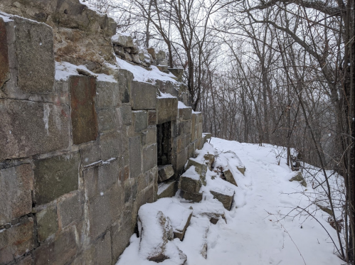 Snow-covered stone ruins surrounded by bare trees on a winter day.
