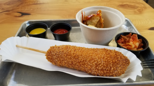 A crispy corn dog on a tray, accompanied by dipping sauces and a cup of kimchi.