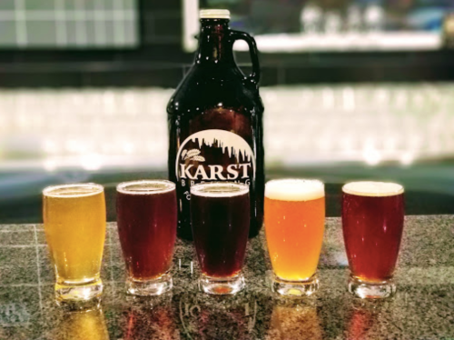 Five glasses of beer in various colors lined up in front of a growler from Karst Brewing Company.